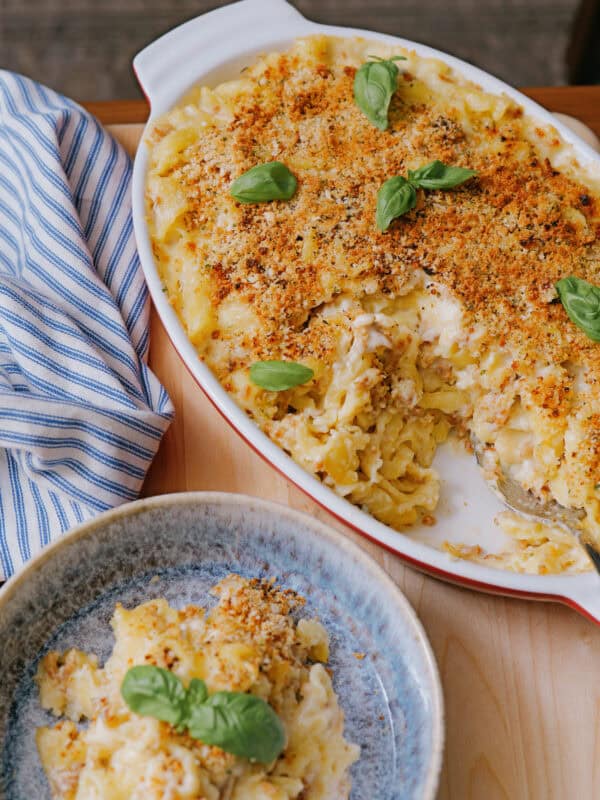 A baked macaroni and cheese casserole topped with golden breadcrumbs and fresh basil leaves, served in a white dish. A portion is served in a blue bowl nearby, with a striped napkin on the side.