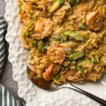 A white, scalloped-edge plate filled with shrimp orzo, asparagus, peas, and grated cheese. A silver spoon rests on the plate, and a green-striped cloth is nearby—perfect for those seeking new shrimp and orzo recipes.