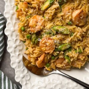 A white, scalloped-edge plate filled with shrimp orzo, asparagus, peas, and grated cheese. A silver spoon rests on the plate, and a green-striped cloth is nearby&mdash;perfect for those seeking new shrimp and orzo recipes.