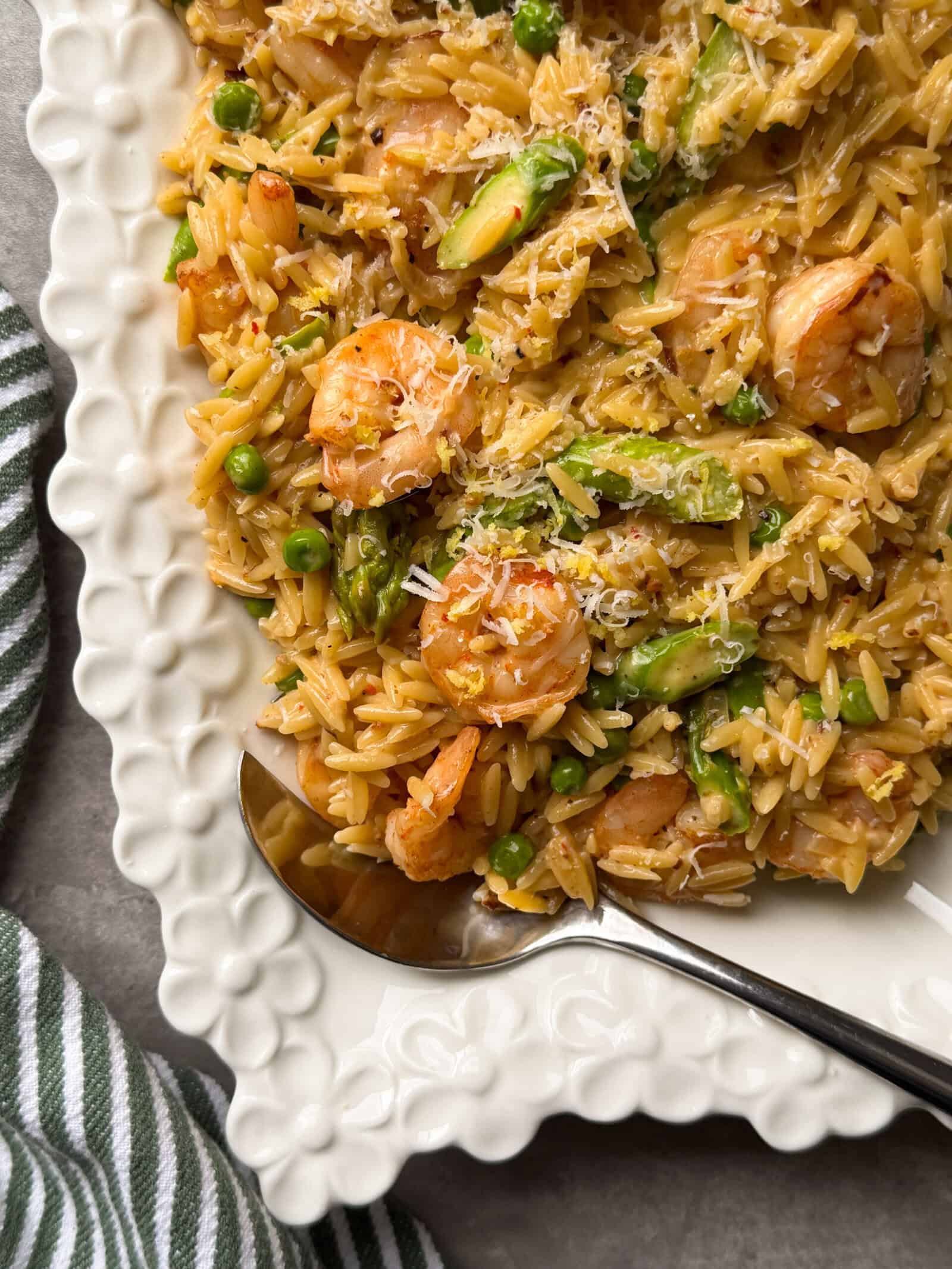 A white, scalloped-edge plate filled with shrimp orzo, asparagus, peas, and grated cheese. A silver spoon rests on the plate, and a green-striped cloth is nearby&mdash;perfect for those seeking new shrimp and orzo recipes.