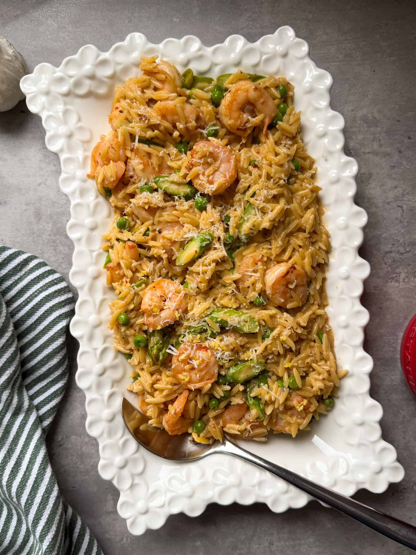 A white rectangular platter with a scalloped edge holds a vibrant shrimp orzo dish mixed with peas and asparagus, garnished with grated cheese. A spoon rests in the dish, and a striped towel is nearby&mdash;perfect for fans of shrimp and orzo recipes.