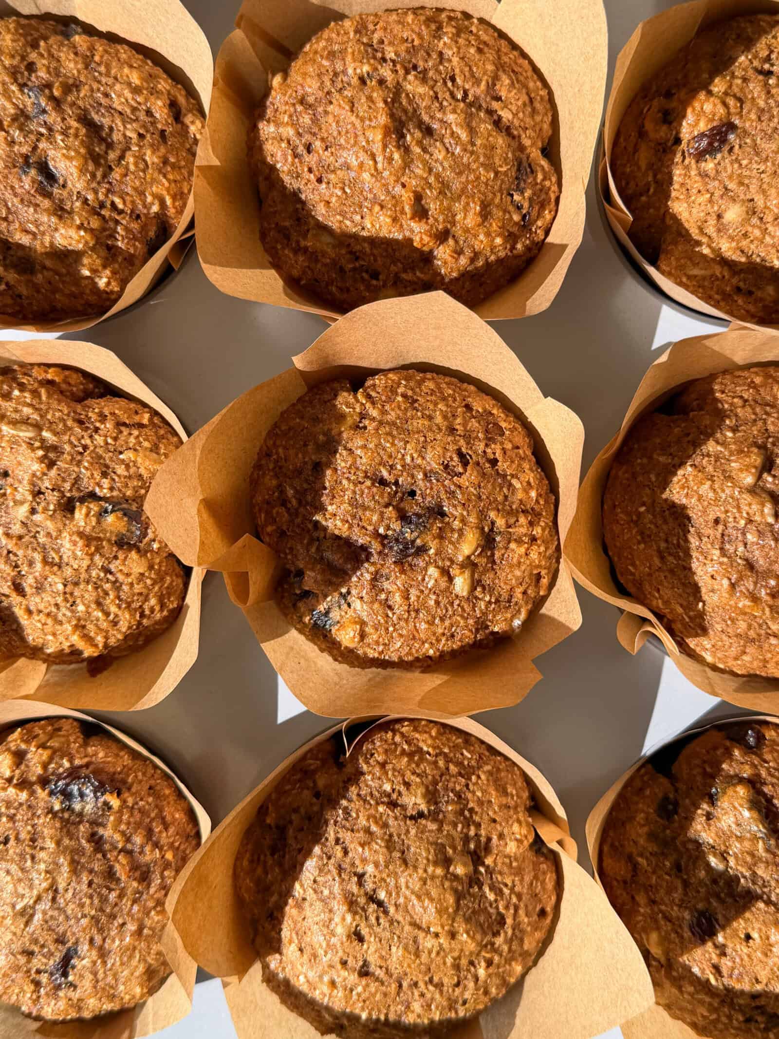 Nine freshly baked bran muffins with a golden-brown top, wrapped in light brown parchment paper liners and arranged in a grid pattern, make the perfect breakfast muffins when lit by bright sunlight.