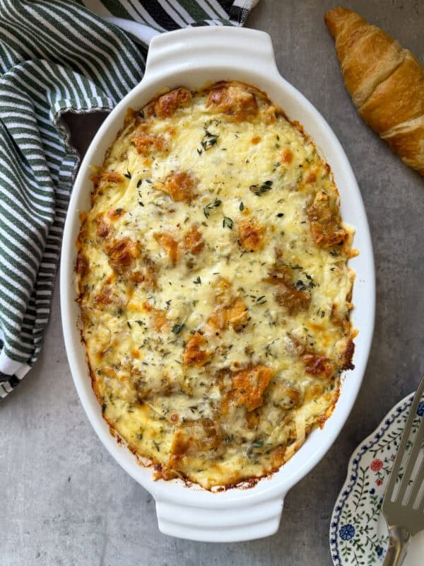 Oval white casserole dish filled with a golden-brown, cheesy croissant breakfast casserole, garnished with herbs. A striped towel, croissant, and floral plate with a serving fork are nearby on a grey surface.