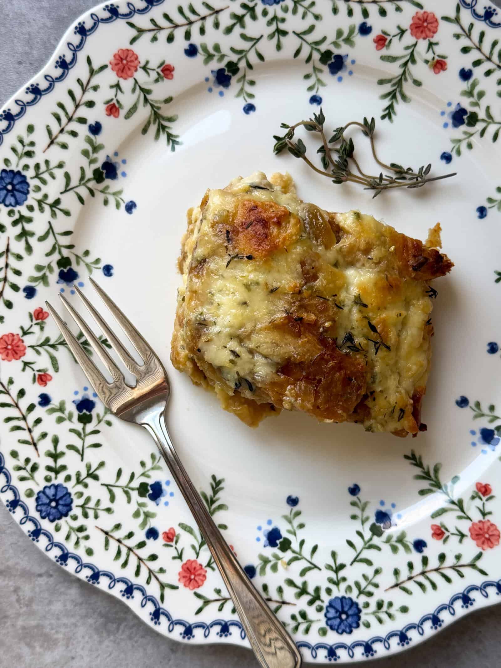 A square piece of savory croissant breakfast casserole garnished with a small sprig of thyme sits on a floral-patterned plate with a silver fork beside it.