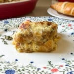 A serving of croissant breakfast casserole with eggs, sausage, and melted cheese sits on a floral-patterned plate. In the background, there’s a croissant and a red baking dish, perfect for trying new breakfast casserole recipes.