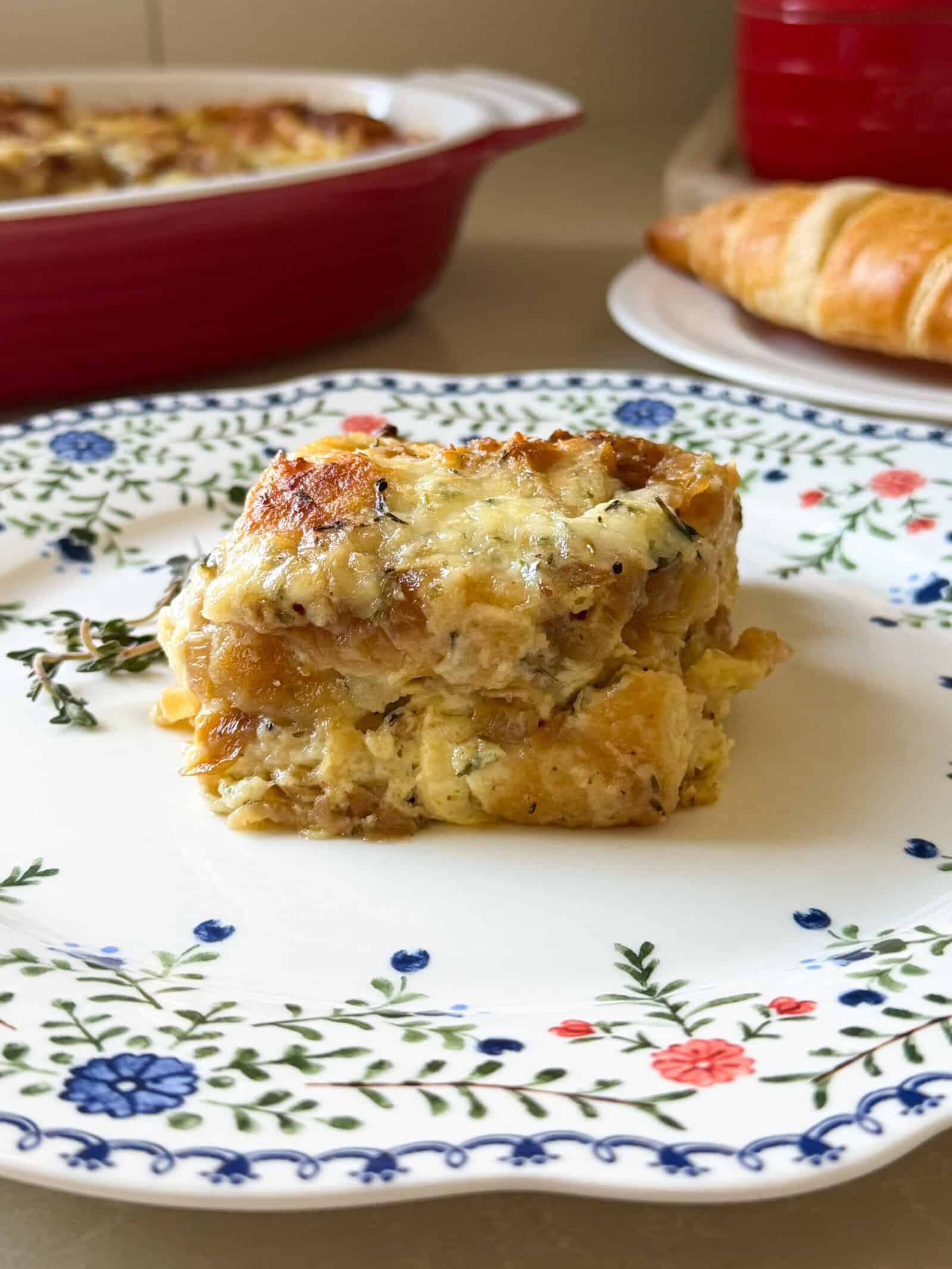 A serving of croissant breakfast casserole with eggs, sausage, and melted cheese sits on a floral-patterned plate. In the background, there’s a croissant and a red baking dish, perfect for trying new breakfast casserole recipes.
