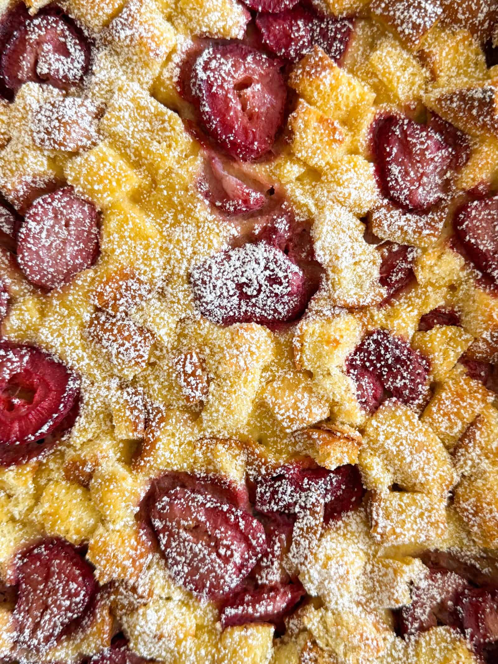 A close-up of a baked dessert with golden bread cubes and slices of red fruit, possibly plums, dusted with powdered sugar—reminiscent of a strawberry french toast casserole fresh from the oven.