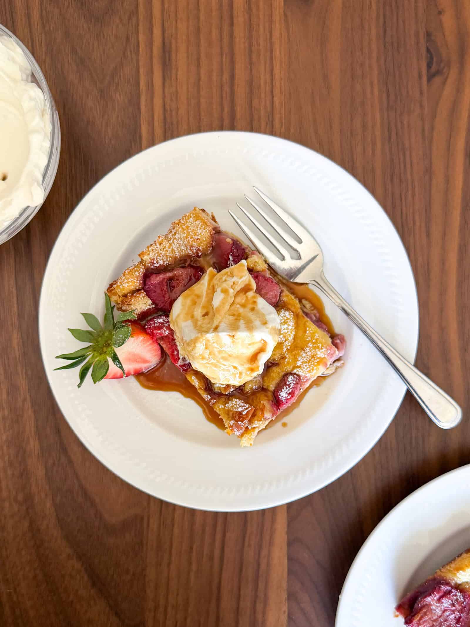 A slice of strawberry french toast casserole, topped with whipped cream and syrup, sits on a white plate with a fork. Fresh strawberries garnish the side, while another plate is partially visible on the wooden table.