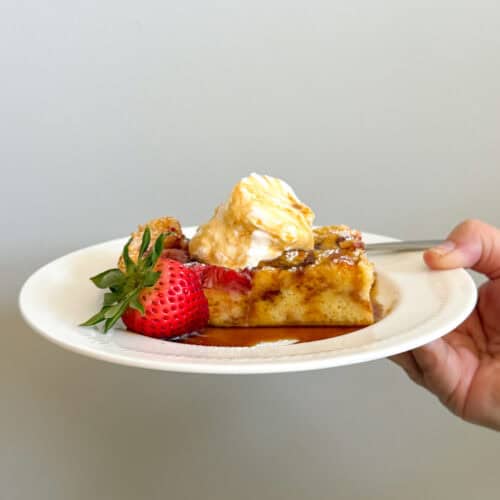 A hand holds a white plate with a slice of strawberry french toast casserole topped with whipped cream and syrup, garnished with a fresh strawberry, against a plain light-colored background.