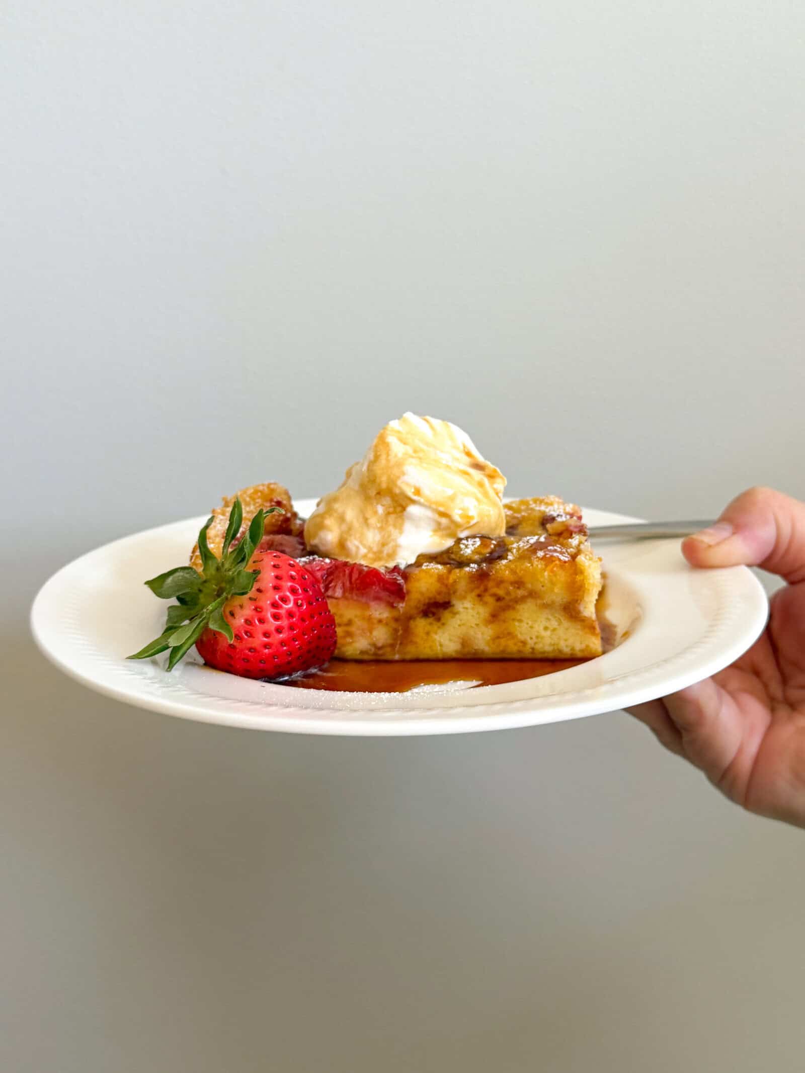 A hand holds a white plate with a slice of strawberry french toast casserole topped with whipped cream and syrup, garnished with a fresh strawberry, against a plain light-colored background.