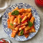 A plate of Rigatoni Arrabbiata with tomato sauce, garnished with fresh basil leaves, is served on a blue floral-patterned dish. A red container, a cloth, and a small plate of red chilies are nearby—classic elements of what is Arrabbiata sauce.