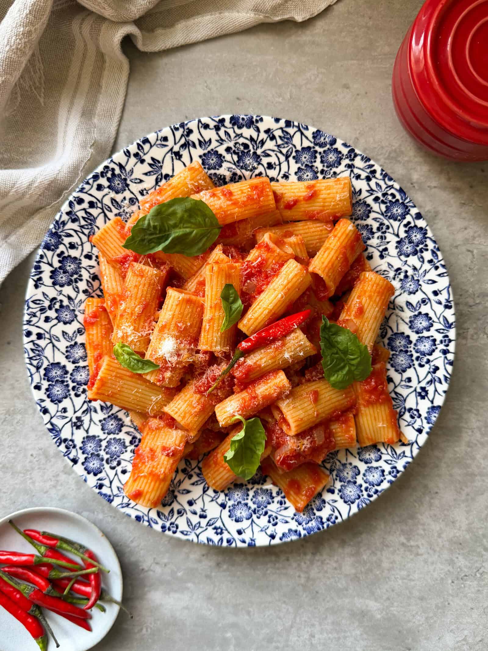 A plate of Rigatoni Arrabbiata with tomato sauce, garnished with fresh basil leaves, is served on a blue floral-patterned dish. A red container, a cloth, and a small plate of red chilies are nearby&mdash;classic elements of what is Arrabbiata sauce.