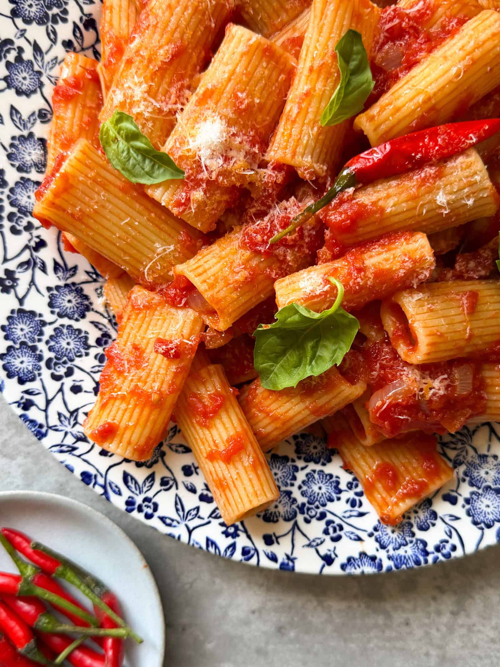 A plate of Rigatoni Arrabbiata in spicy tomato sauce, garnished with fresh basil and red chili peppers, served on a blue and white floral-patterned dish. A small plate of red and green chilies sits nearby.
