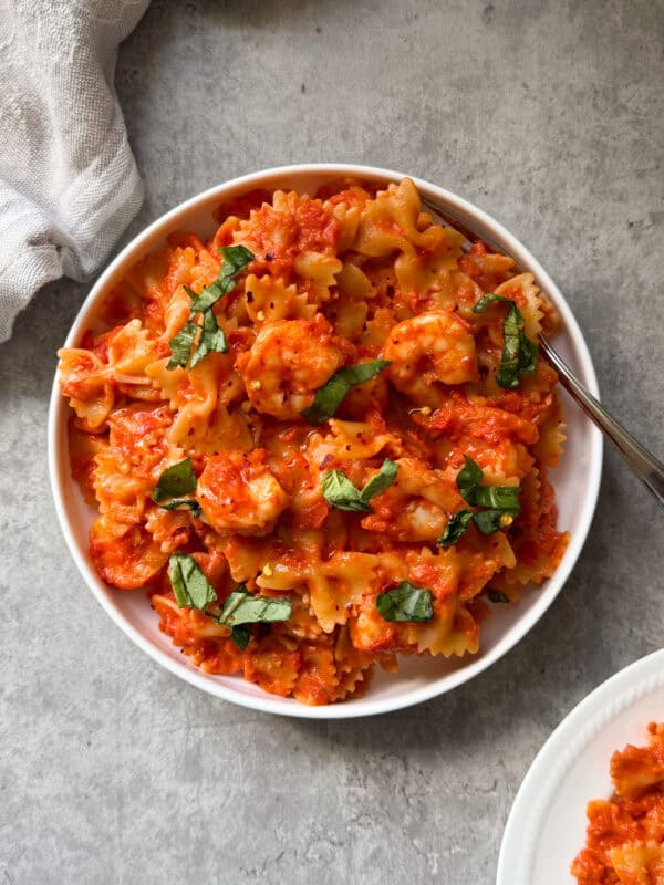 A bowl of Bowtie Pasta with Shrimp & Creamy Rosè sauce, topped with fresh basil, sits on a gray surface beside a spoon and a white napkin, making for an elegant and delicious meal.