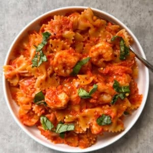 A white bowl filled with Bowtie Pasta with Shrimp & Creamy Rosè, garnished with chopped fresh basil in a velvety rose sauce, sits on a light gray surface.
