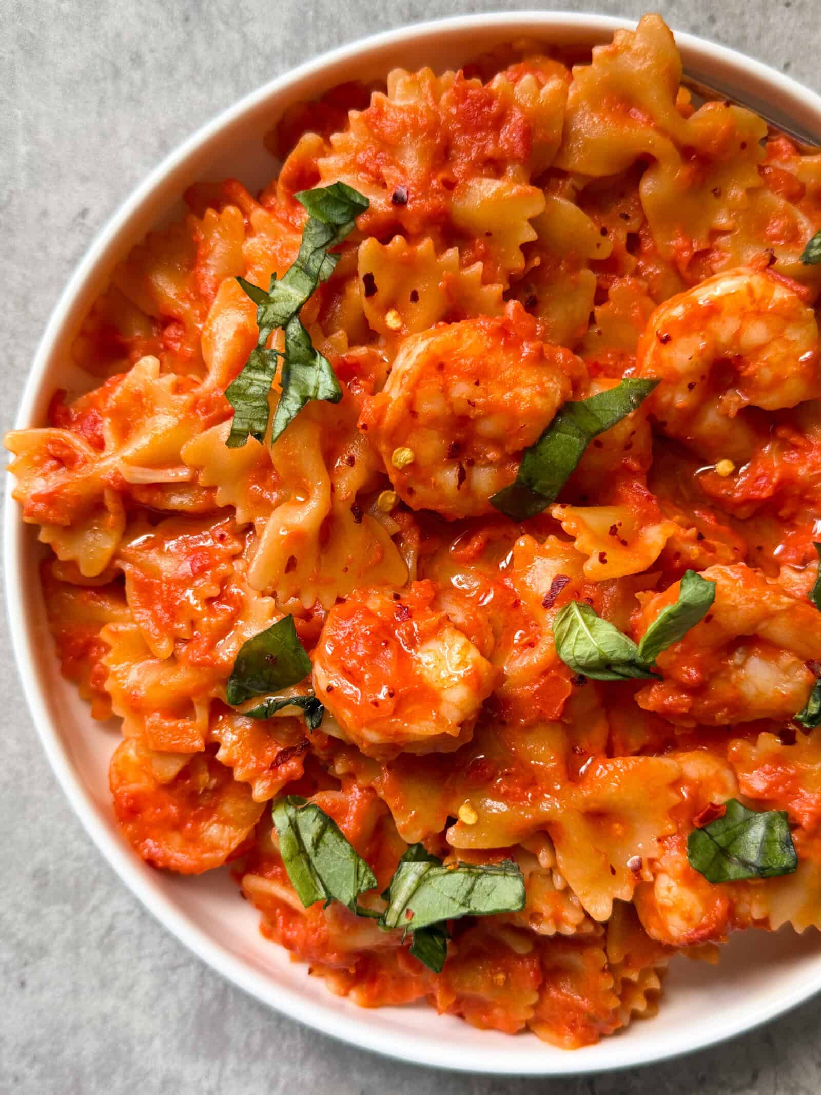 A bowl of Bowtie Pasta with Shrimp & Creamy Ros&egrave; sauce, garnished with fresh basil leaves, sits on a light-colored surface. This creamy shrimp pasta is the perfect blend of flavors and elegance.