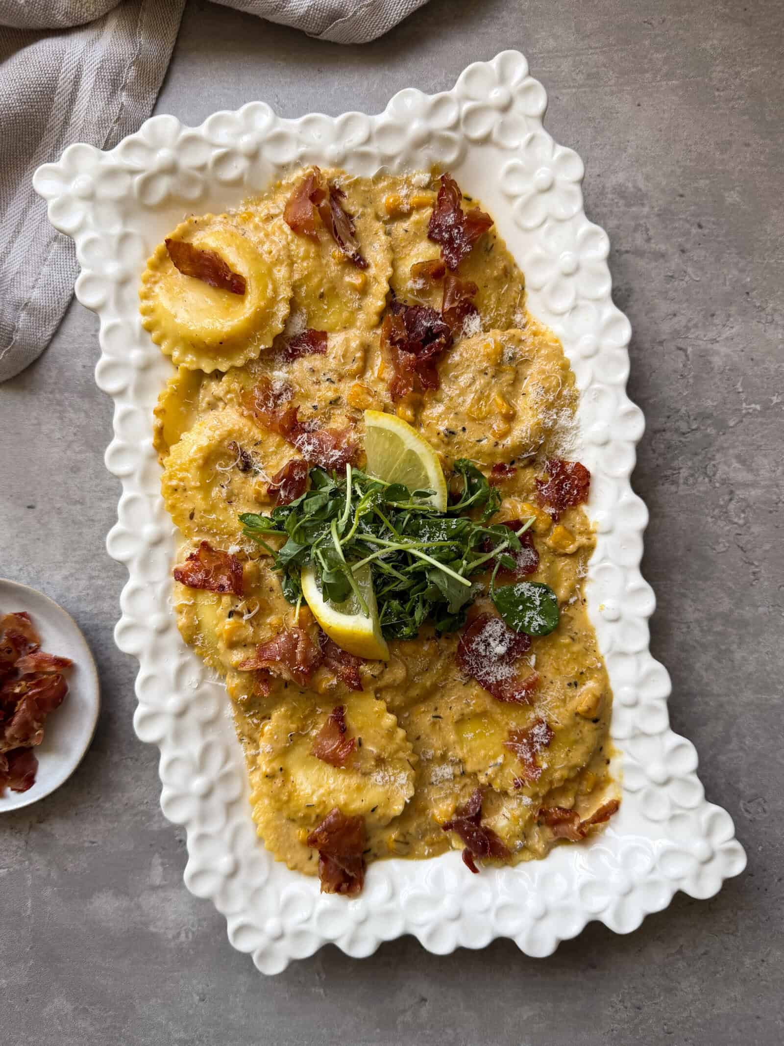 A decorative white plate features a creamy yellow ravioli recipe, topped with crispy prosciutto, grated cheese, fresh arugula, and lemon wedges&mdash;perfect inspiration for your next frozen ravioli recipes. A small dish of prosciutto sits nearby on a gray surface.