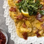 A white scalloped plate of ravioli in a creamy sauce, topped with crispy prosciutto, grated cheese, fresh arugula, and lemon wedges—a delicious take on classic ravioli recipes. A small side dish with more crispy prosciutto is visible on the left.