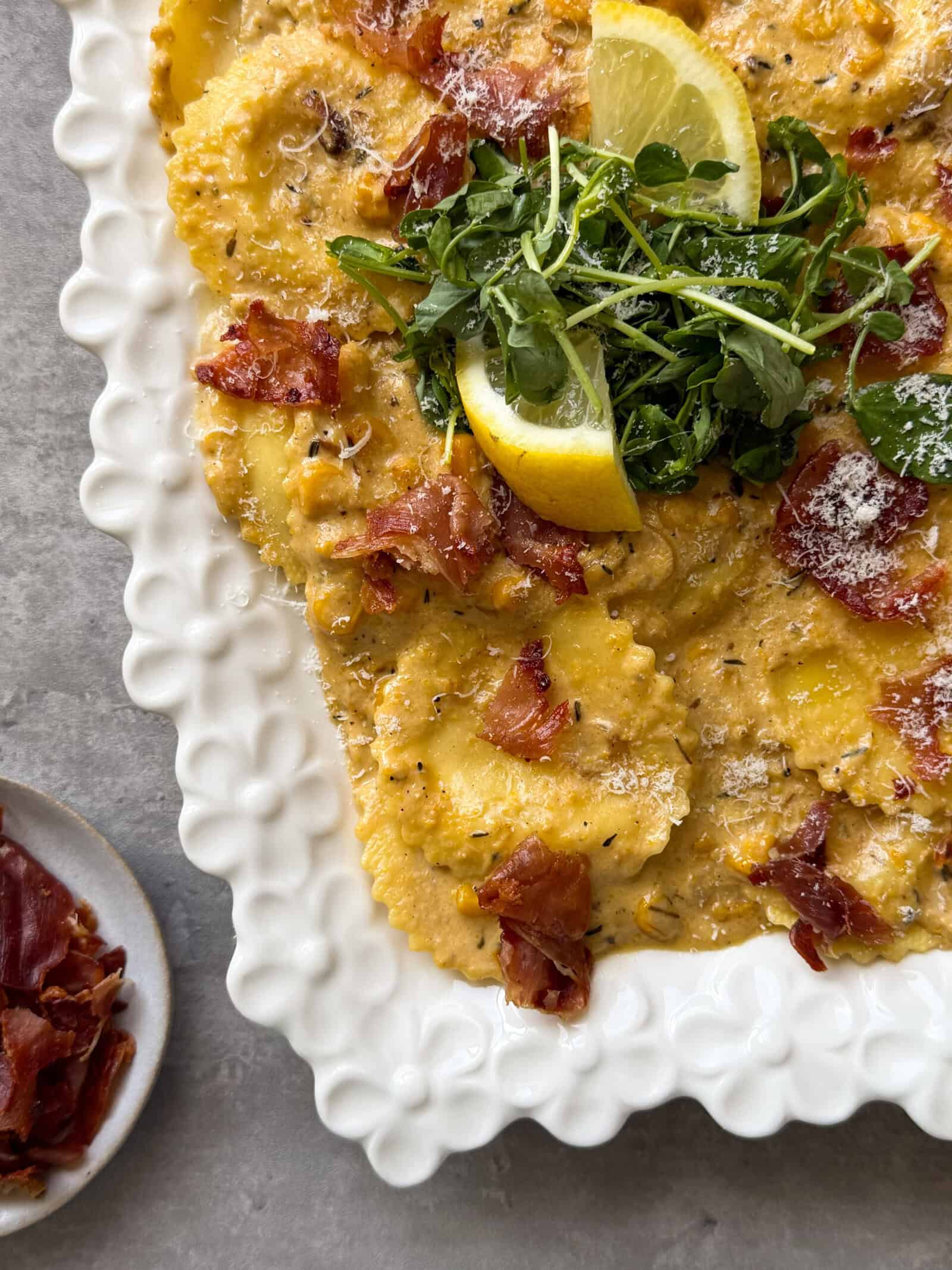 A white scalloped plate of ravioli in a creamy sauce, topped with crispy prosciutto, grated cheese, fresh arugula, and lemon wedges—a delicious take on classic ravioli recipes. A small side dish with more crispy prosciutto is visible on the left.