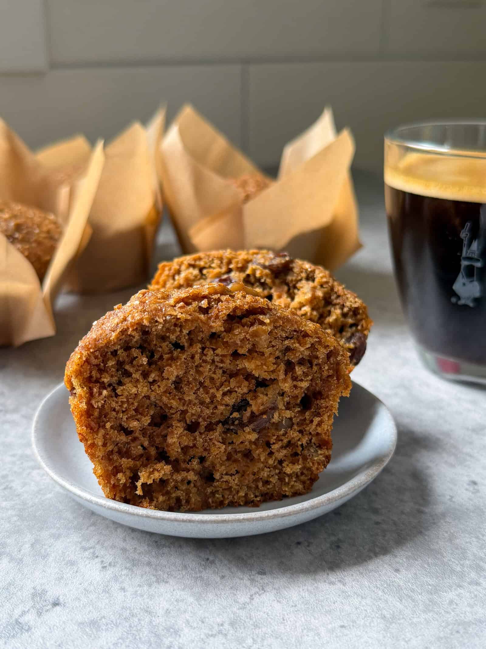A sliced raisin bran muffin sits on a small plate next to a glass of dark coffee, with more bran muffins in brown paper wrappers in the background on a light countertop.