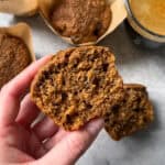 A hand holds half of a moist bran muffin, showing its texture. Other whole bran muffins in brown paper liners and a cup of coffee are on a light surface in the background.