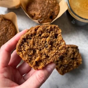 A hand holds half of a moist bran muffin, showing its texture. Other whole bran muffins in brown paper liners and a cup of coffee are on a light surface in the background.