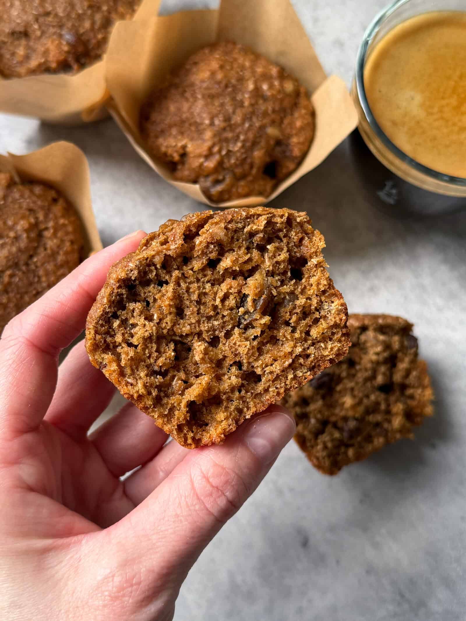 A hand holds half of a moist bran muffin, showing its texture. Other whole bran muffins in brown paper liners and a cup of coffee are on a light surface in the background.
