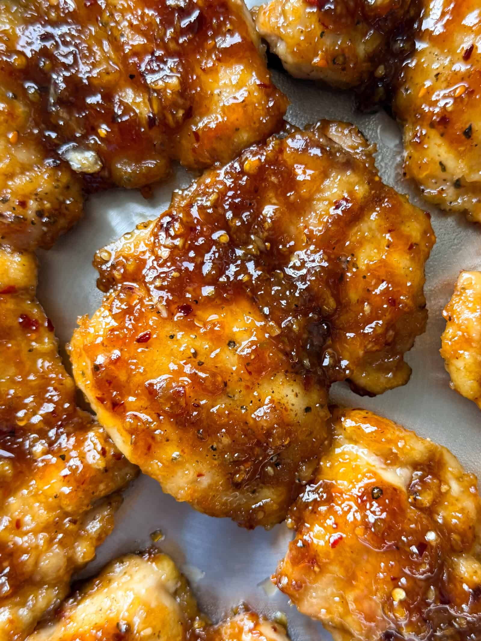Close-up of crispy, golden-brown honey glazed chicken thighs coated in a sticky, glossy sauce with visible black pepper and seasoning, arranged on a metallic surface—perfect inspiration for your next chicken thighs recipes.