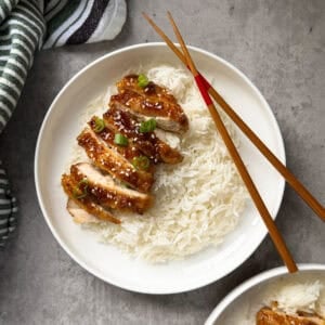 A plate of white rice topped with sliced honey glazed chicken, garnished with chopped green onions and sesame seeds. Wooden chopsticks rest on the edge of the plate—a delicious choice for those seeking chicken thighs recipes. A striped cloth is visible to the side.