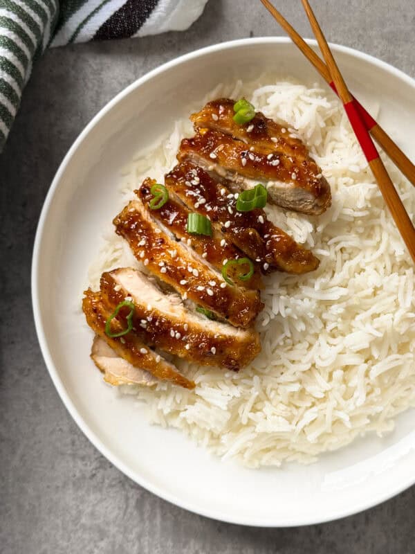 A white plate with sliced honey glazed chicken topped with sesame seeds and chopped green onions, served over white rice. Perfect for those seeking easy chicken thighs recipes. Chopsticks rest on the side of the plate.