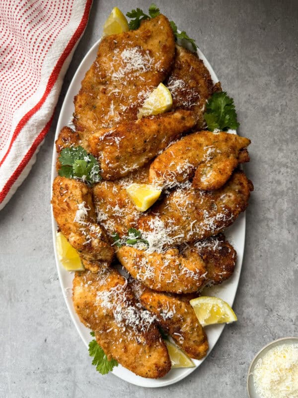 A white oval platter with golden-brown Caesar Chicken Cutlets, garnished with grated cheese, parsley, and lemon wedges, set on a gray surface beside a red striped towel.