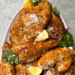 A plate of golden-brown Chicken Cutlets, garnished with grated cheese, fresh cilantro, and lemon wedges, set on a light gray surface—an inviting Chicken Cutlets Recipe perfect for any meal.