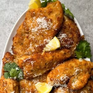 A plate of golden-brown Chicken Cutlets, garnished with grated cheese, fresh cilantro, and lemon wedges, set on a light gray surface—an inviting Chicken Cutlets Recipe perfect for any meal.