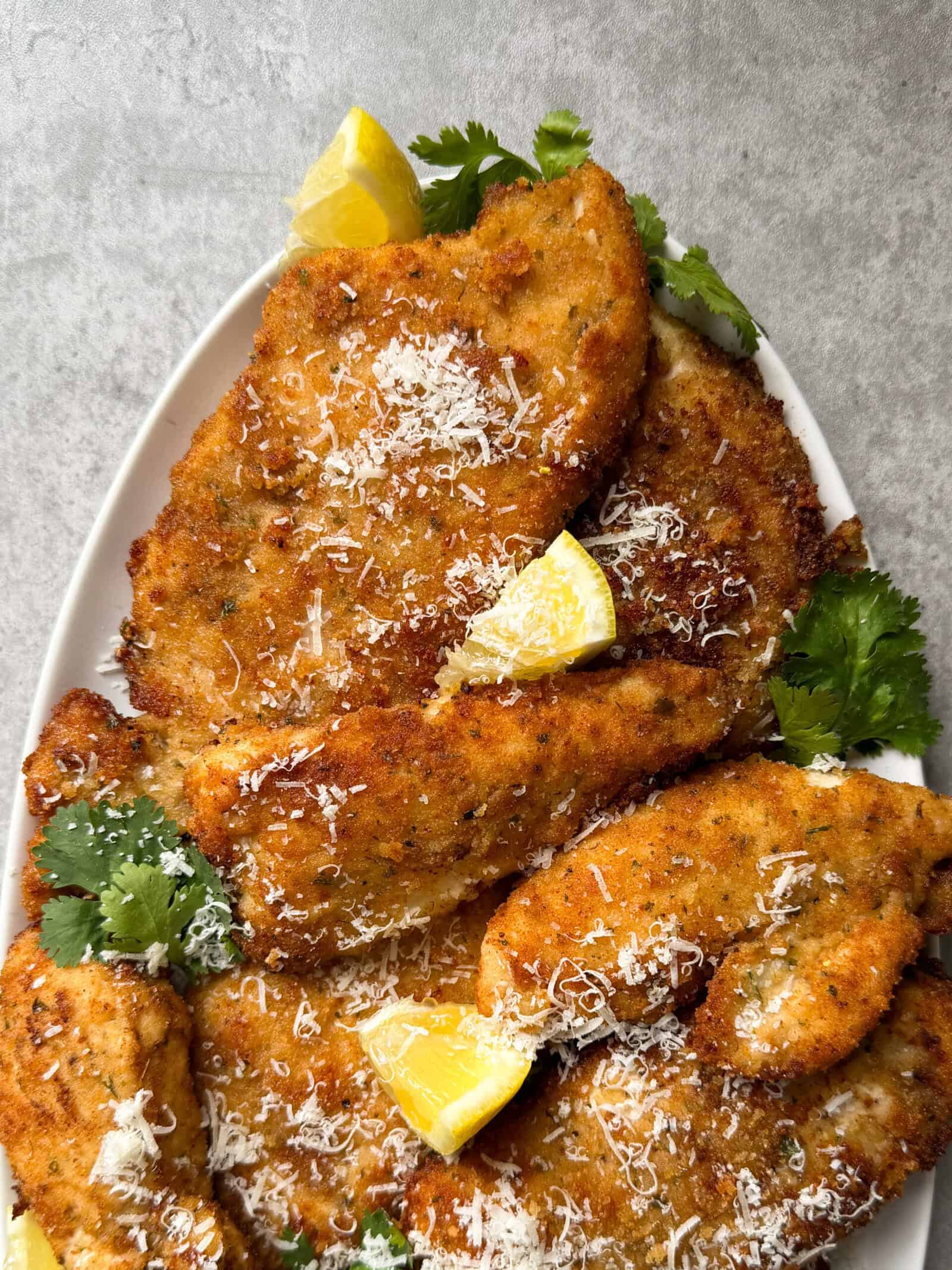 A plate of golden-brown Chicken Cutlets, garnished with grated cheese, fresh cilantro, and lemon wedges, set on a light gray surface—an inviting Chicken Cutlets Recipe perfect for any meal.