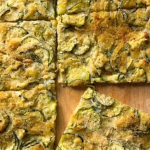 Golden, rectangular slices of Zucchini Flatbread on a wooden surface. This zucchini flatbread recipe features thin zucchini slices and is sprinkled with herbs and parmesan, with one piece slightly separated from the others.