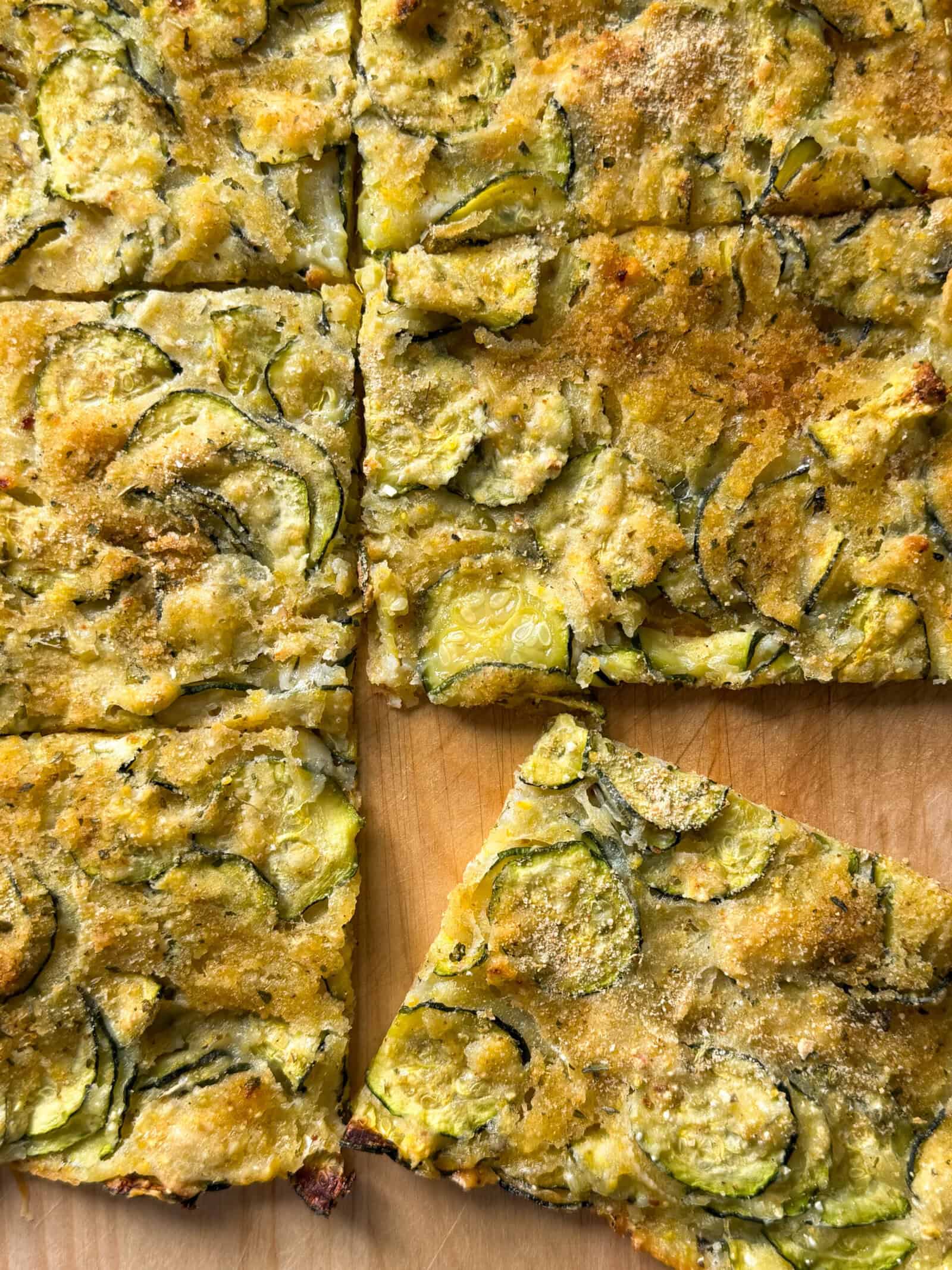 Golden, rectangular slices of Zucchini Flatbread on a wooden surface. This zucchini flatbread recipe features thin zucchini slices and is sprinkled with herbs and parmesan, with one piece slightly separated from the others.