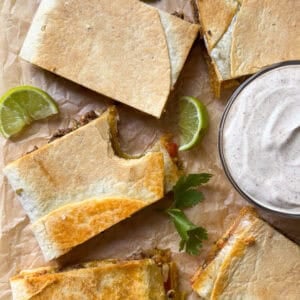 Rectangular quesadilla slices on parchment paper with lime wedges and cilantro leaves, next to a bowl of creamy dipping sauce. One quesadilla has a bite taken out of it—perfect for fans of sheet pan recipes or homemade crunchwrap creations.