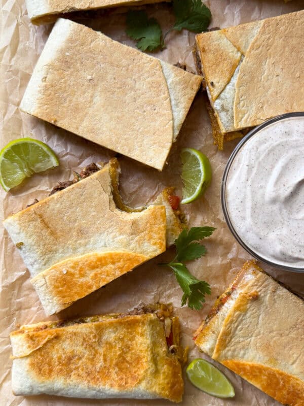Rectangular quesadilla slices on parchment paper with lime wedges and cilantro leaves, next to a bowl of creamy dipping sauce. One quesadilla has a bite taken out of it—perfect for fans of sheet pan recipes or homemade crunchwrap creations.