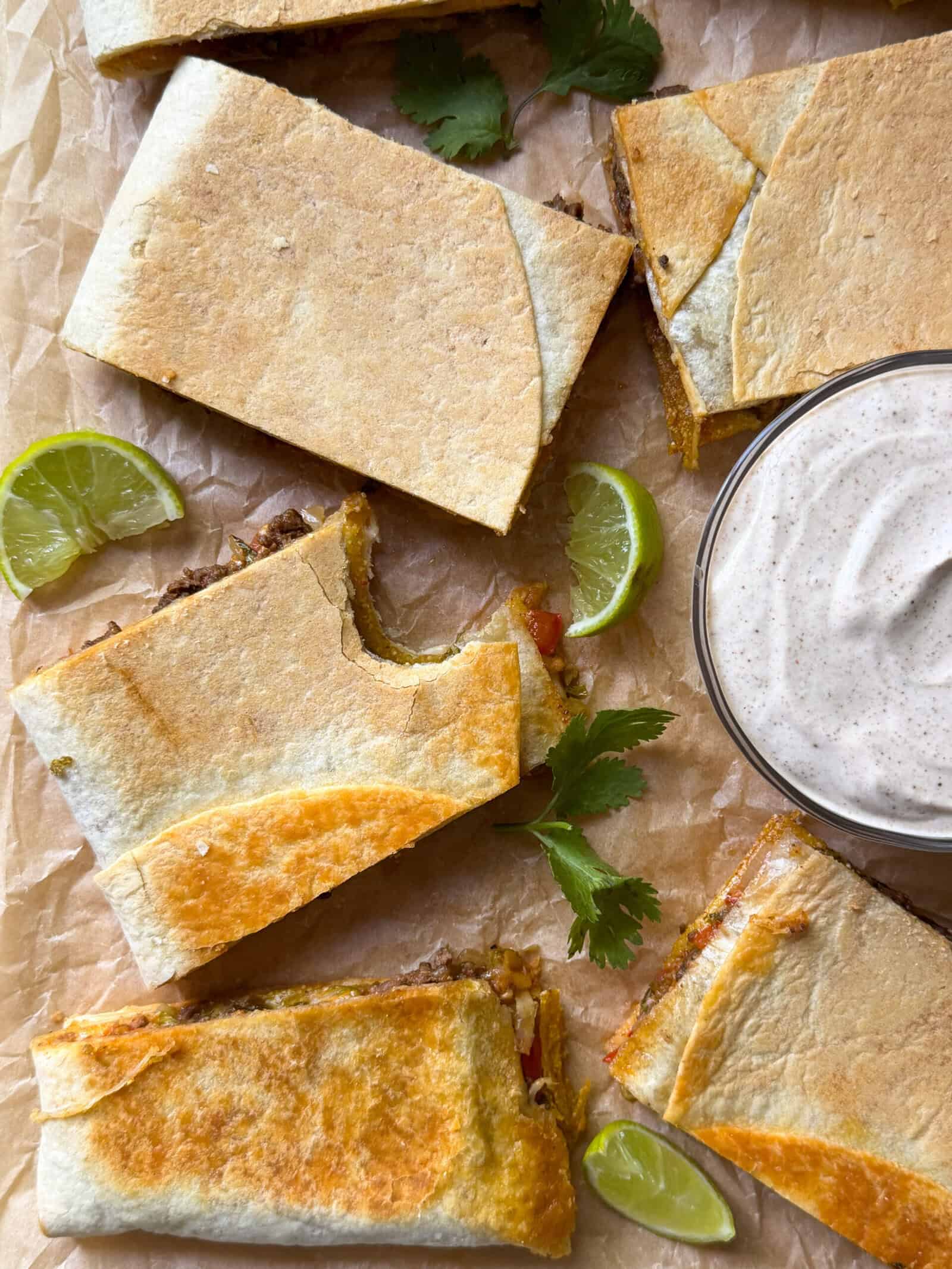 Rectangular quesadilla slices on parchment paper with lime wedges and cilantro leaves, next to a bowl of creamy dipping sauce. One quesadilla has a bite taken out of it—perfect for fans of sheet pan recipes or homemade crunchwrap creations.