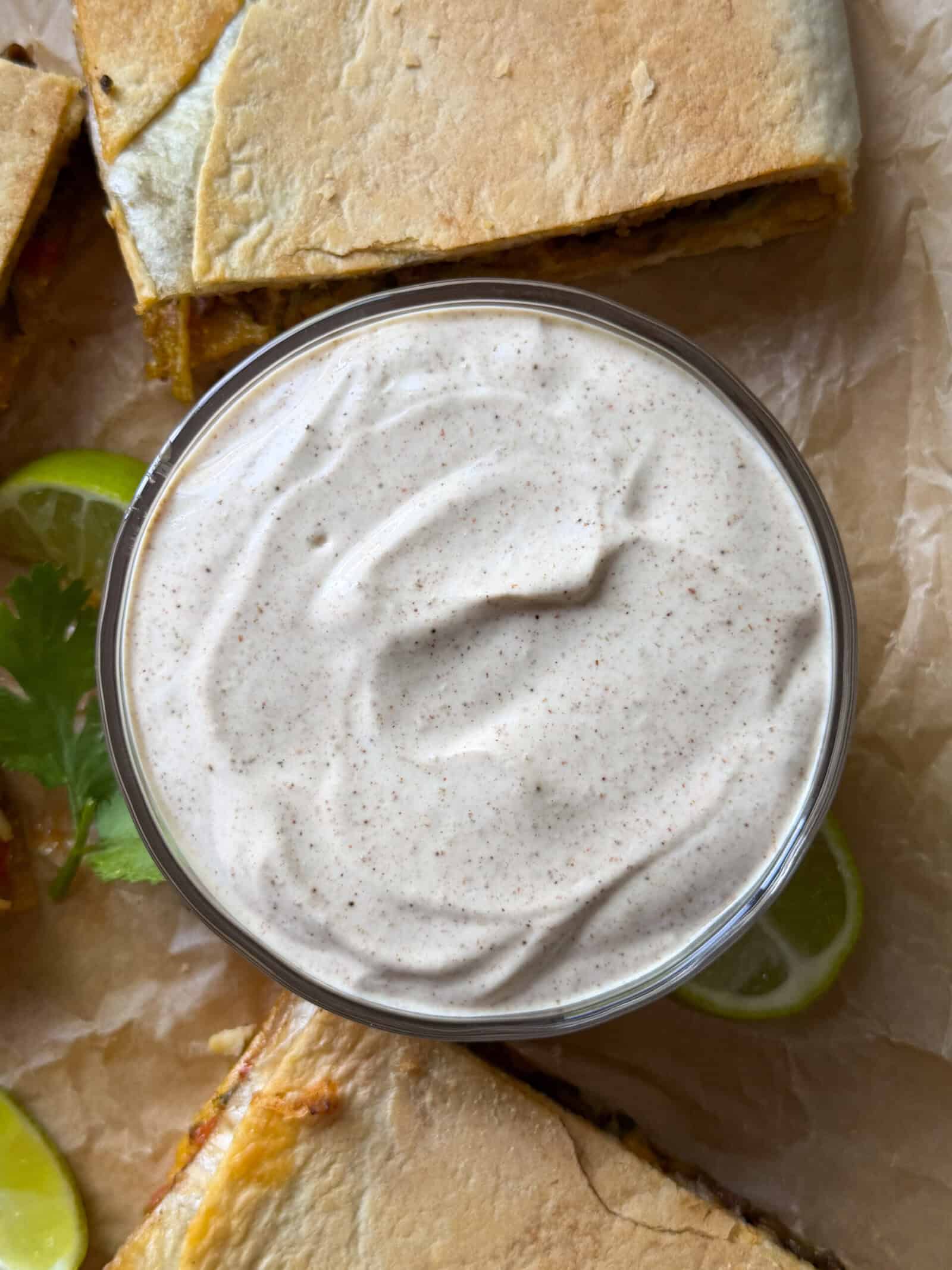 A bowl of creamy, light tan dipping sauce with a sprinkle of spices sits on parchment paper, surrounded by cut sheet pan crunchwraps, lime slices, and a sprig of cilantro—perfect for your next homemade crunchwrap night.