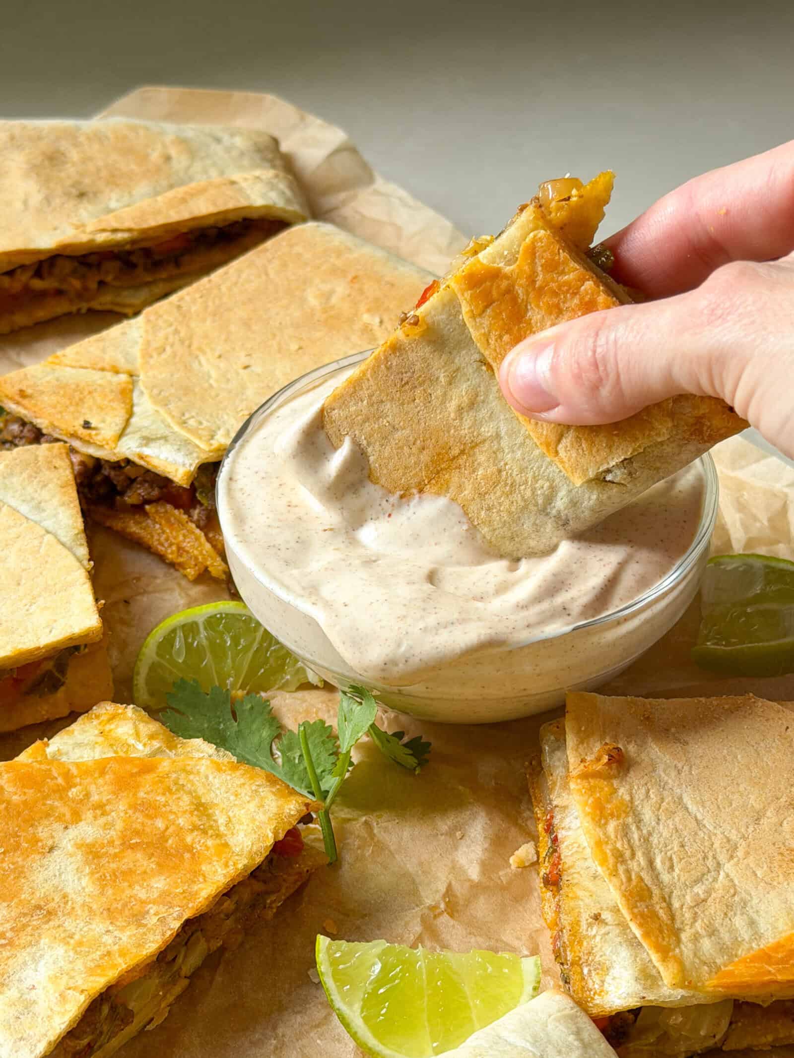 A hand dips a triangular sheet pan crunchwrap slice into a bowl of creamy sauce. Other crunchwrap pieces, lime wedges, and cilantro are arranged on parchment paper nearby—perfect inspiration for easy sheet pan recipes.