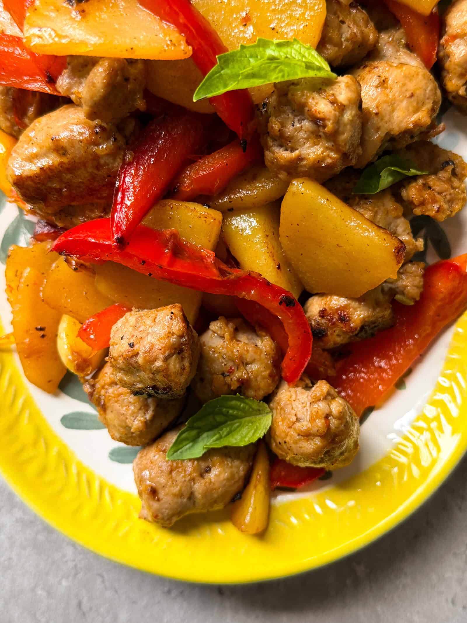 A close-up of a plate featuring a delicious one skillet fried sausage potatoes & peppers recipe, with cooked sausage pieces, sliced yellow potatoes, and red bell peppers, garnished with fresh basil on a colorful yellow-rimmed plate.