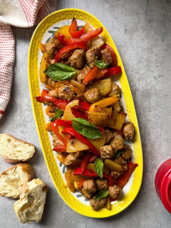 An oval yellow platter featuring a vibrant one skillet fried sausage potatoes & peppers mix, with fresh basil, sits on a gray surface beside pieces of bread and a red towel.