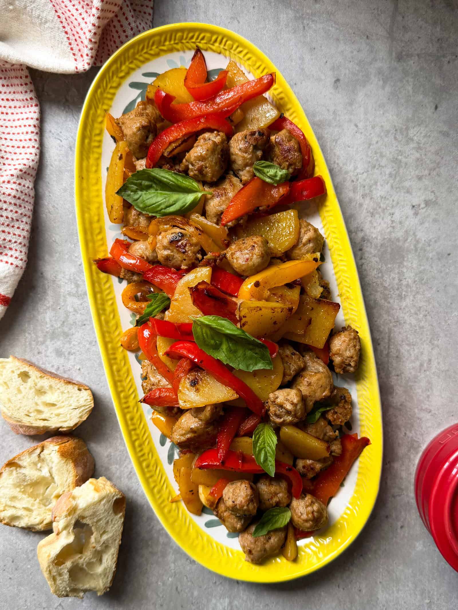 An oval yellow platter featuring a vibrant one skillet fried sausage potatoes & peppers mix, with fresh basil, sits on a gray surface beside pieces of bread and a red towel.