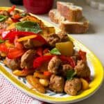 A yellow-edged platter holds sautéed sausage potatoes and peppers with onions and fresh basil leaves. Slices of bread and a red pot sit in the background on a countertop, with a striped kitchen towel nearby.