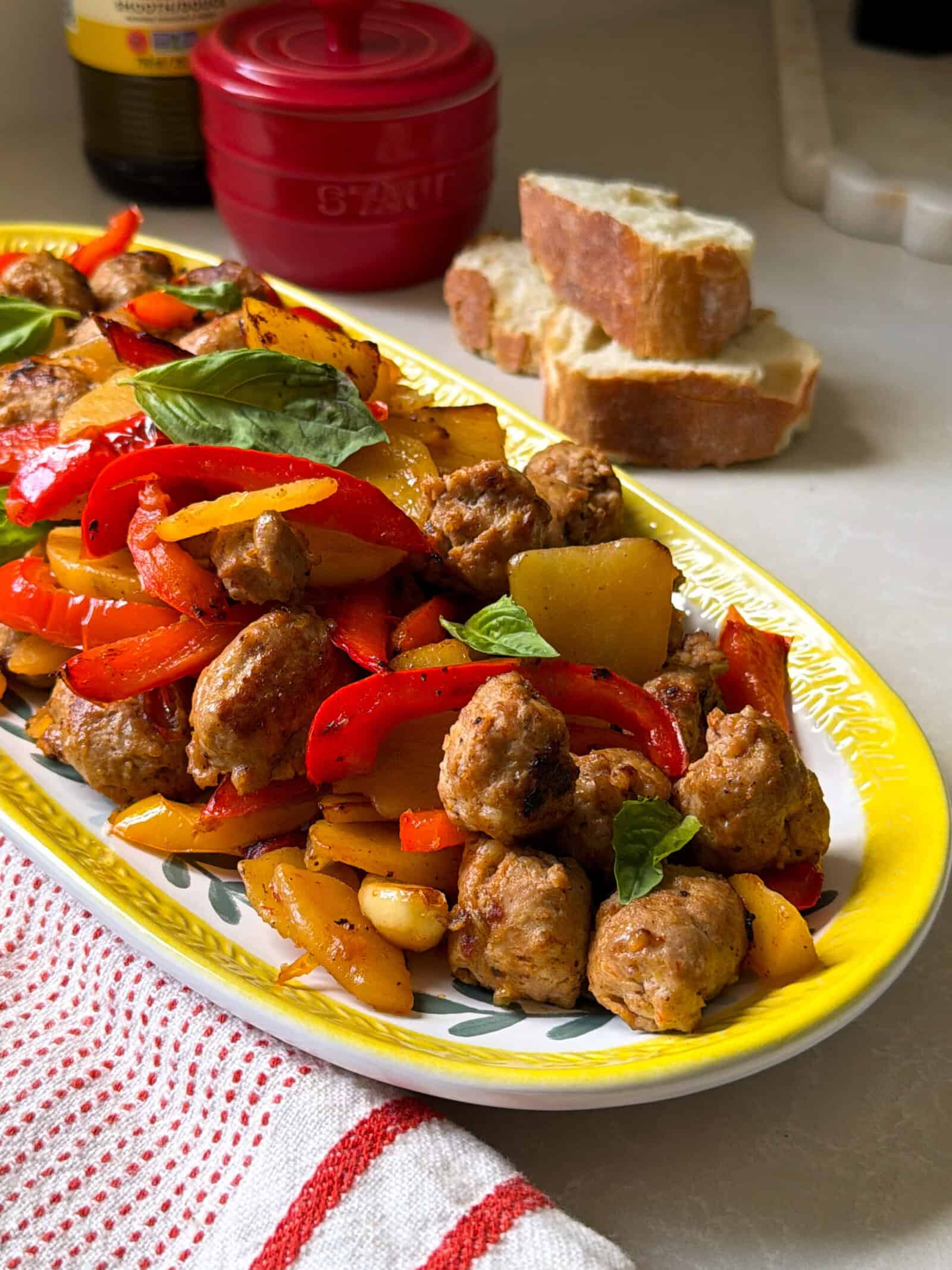 A yellow-edged platter holds sautéed sausage potatoes and peppers with onions and fresh basil leaves. Slices of bread and a red pot sit in the background on a countertop, with a striped kitchen towel nearby.
