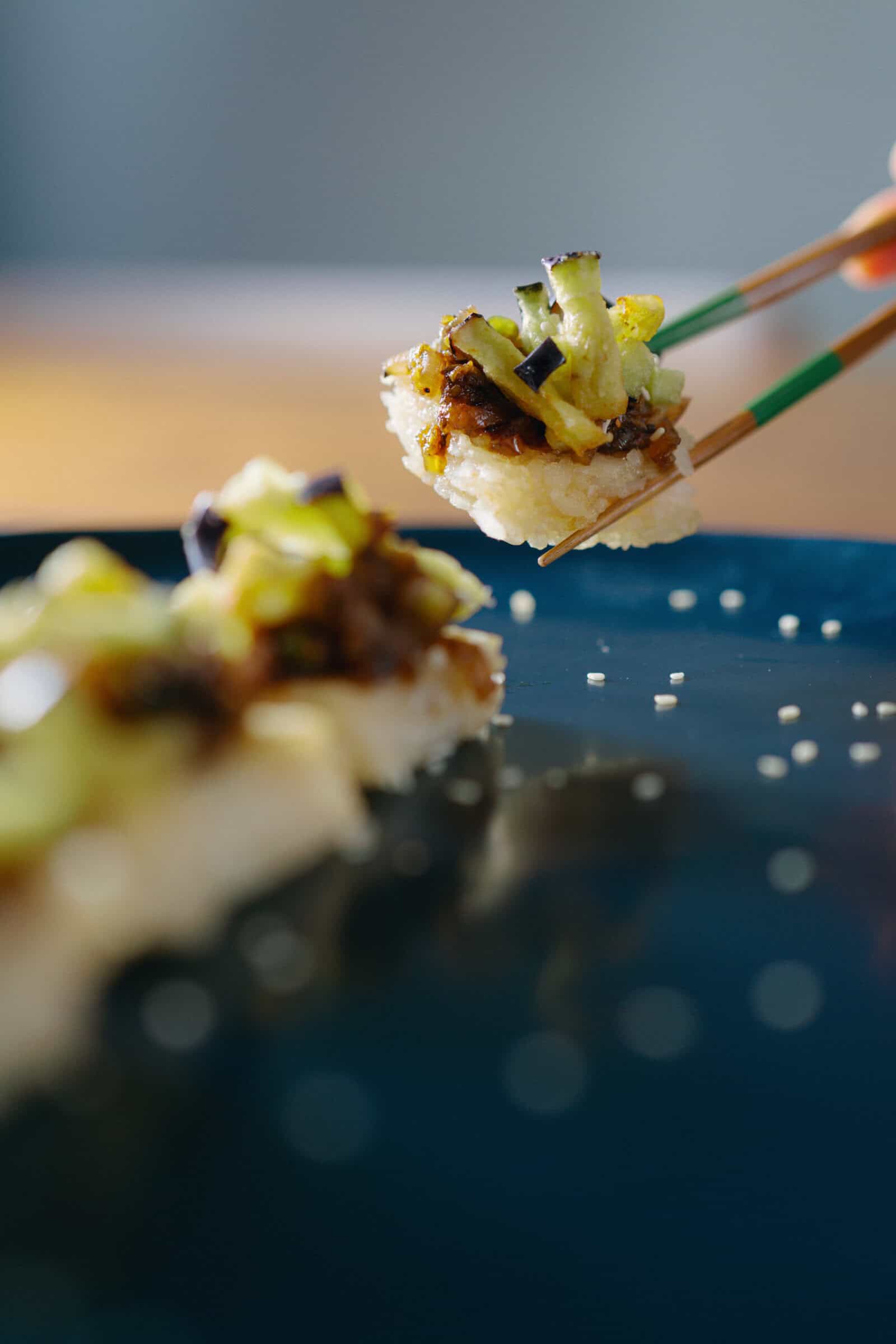 Close-up of chopsticks holding a piece of sushi topped with chopped vegetables over a dark plate, with another piece of sushi and scattered rice grains visible in the background.