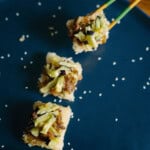 Four pieces of sushi with shredded vegetables are on a dark blue plate sprinkled with sesame seeds; a hand is using chopsticks to pick up one piece.