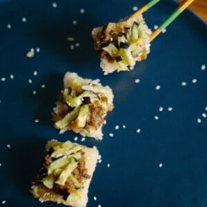 Four pieces of sushi with shredded vegetables are on a dark blue plate sprinkled with sesame seeds; a hand is using chopsticks to pick up one piece.