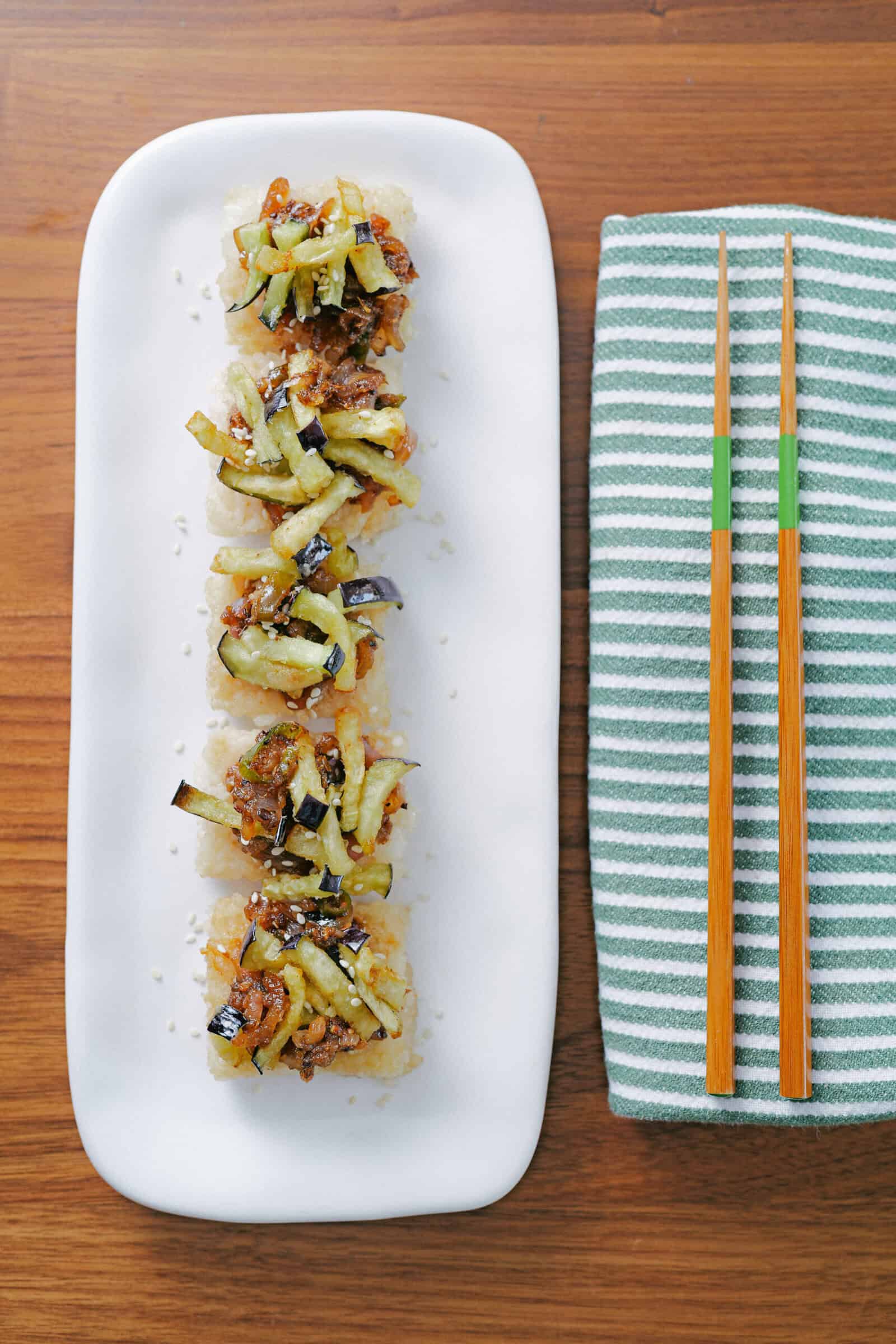 A rectangular white plate holds five pieces of sushi topped with shredded vegetables, next to a folded green and white striped napkin and a pair of wooden chopsticks with green accents on a wooden table.